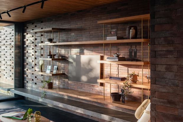 Warm contemporary interior featuring a full-height dark brick feature wall with integrated timber shelving and metal supports. The shelving displays ceramics, books, framed artwork and plants, with a long built-in bench below. A timber-lined ceiling, track lighting, concrete-toned surfaces and large glazing create an industrial yet refined living space.