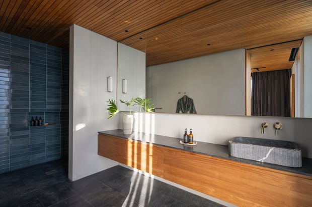 Contemporary bathroom featuring a long timber vanity with dark stone benchtop, vessel basin and wall-mounted brass tapware. A dark blue-grey tiled shower area is visible to the left, while a timber-lined ceiling, pale plaster walls, integrated storage and warm natural light create a refined, modern atmosphere.