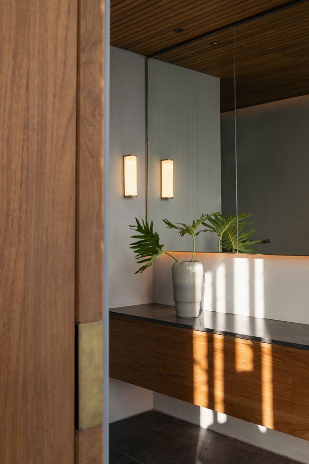 A refined interior vignette viewed through a timber-framed opening, featuring a dark timber slatted ceiling, pale plaster walls, a dark stone benchtop, and warm timber cabinetry or a built-in console. Two vertical wall sconces illuminate the space, while a ceramic vase with leafy greenery adds a natural accent. Strong sunlight creates dramatic shadows across the cabinetry and floor.
