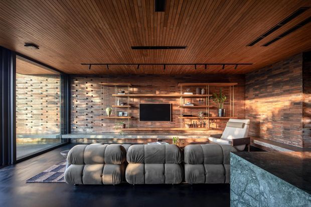 A warm, modern living room with a low modular sectional sofa facing a wall-mounted television and open timber shelving. The room features extensive horizontal timber wall cladding, a slatted timber ceiling, dark flooring, large corner glazing, and a dark stone-clad kitchen or bar counter in the foreground.