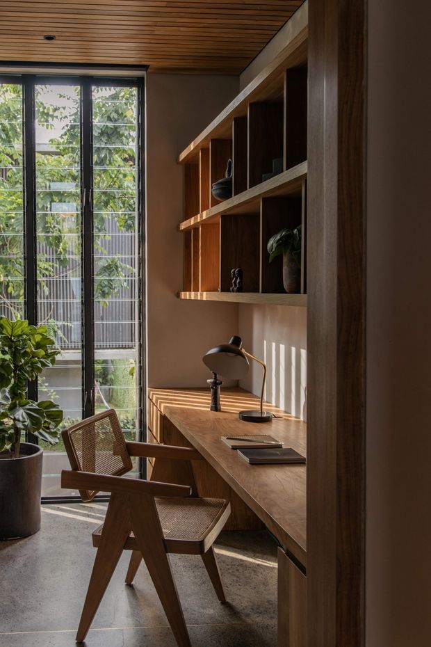 A warm, compact home office with a built-in timber desk and open shelving, a woven timber armchair, and a black desk lamp. Full-height glazed doors or windows bring in strong natural light, while the timber slatted ceiling, concrete floor, plants, and deep wood joinery create a calm, natural atmosphere.