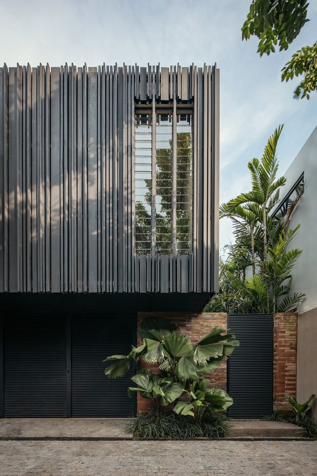 Street-facing contemporary building facade with dark horizontal garage doors, a brick boundary wall, and a prominent upper-level screen of closely spaced vertical metal fins. A tall glazed opening is set within the fin screen, while tropical planting softens the frontage. The image is photographed from outside at street level.