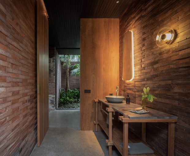 Warm, moody bathroom interior with exposed reddish brick walls, dark horizontal timber wall cladding, a timber ceiling, polished concrete flooring, and a timber vanity with a vessel basin. A large vertical mirror and sculptural wall lighting sit above the vanity, while an open timber door leads toward a planted courtyard or exterior passage.