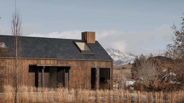 A single-storey dark timber-clad house set in a dry mountainous landscape. The building has a broad pitched roof covered in dark shingles, a prominent timber-clad chimney, and a roof skylight. Large dark-framed glazed openings and a covered entry sit beneath the eaves. Sparse deciduous trees and golden grasses surround the house, with snow-capped mountains visible behind it.
