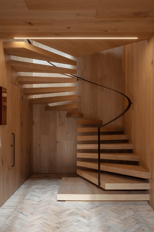 A sculptural open-riser staircase occupies a compact enclosed stair hall. Warm timber panels clad the walls, ceiling, stair treads, and landing, while a slender dark metal handrail curves around the switchback stair. Narrow glazed openings bring daylight between the floating timber steps, and the floor is finished in a pale herringbone pattern.