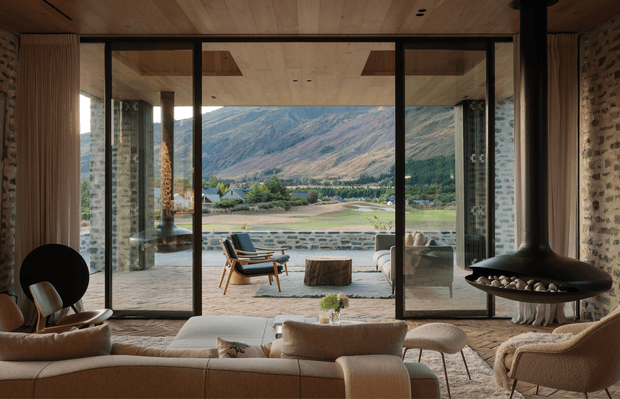 A warm, mid-century-inspired living room with a low contemporary profile, exposed brick walls, timber-lined ceiling, and expansive black-framed sliding glass doors opening to a stone terrace and dramatic mountain landscape. The room features a large cream sectional sofa, sculptural lounge chairs, a suspended black fireplace, and natural timber and stone finishes.