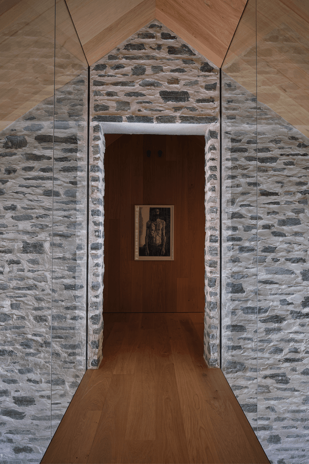 A dramatic architectural interior showing a narrow doorway set within a rough exposed-stone wall. The opening leads into a warm, dark timber-lined room with matching timber flooring, while large floor-to-ceiling mirrored panels flank the stone wall and reflect the space. A small framed artwork hangs on the rear timber wall.