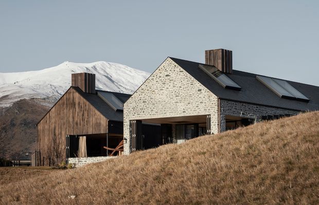 Exterior view of a contemporary alpine residence on a sloping grassy site, with a steep dark pitched roof, pale irregular stone masonry gable, vertical timber-clad volumes, roof windows, and prominent timber-clad chimneys. Snow-covered mountains form the background.