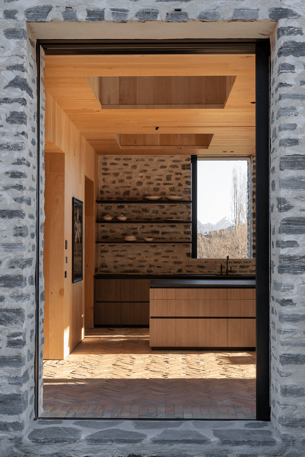 View through a dark-framed doorway into a warm, compact contemporary kitchen. The space features extensive natural timber lining on the walls and ceiling, recessed ceiling openings, a dark stone or engineered-stone benchtop, timber cabinetry, exposed irregular stone masonry on the rear wall, open display shelving, and a herringbone brick floor. A tall window brings in strong daylight and frames a mountainous landscape beyond.