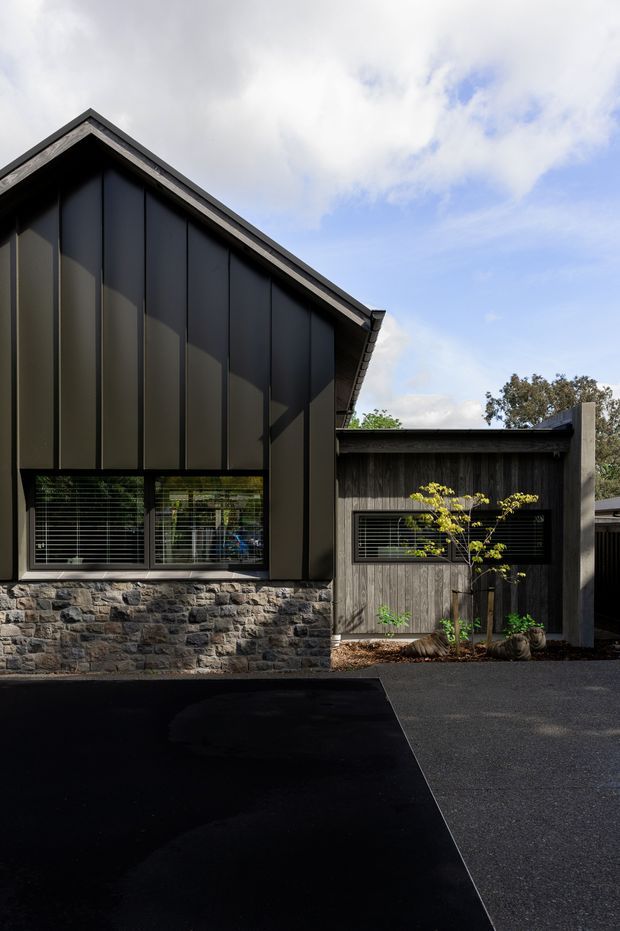 Exterior view of a contemporary gabled building with dark vertical metal cladding, a rough natural-stone base, timber-clad adjoining wing, horizontal window blinds, and a dark paved forecourt. The strongly pitched roof and contrasting materials create a restrained, modern architectural composition.
