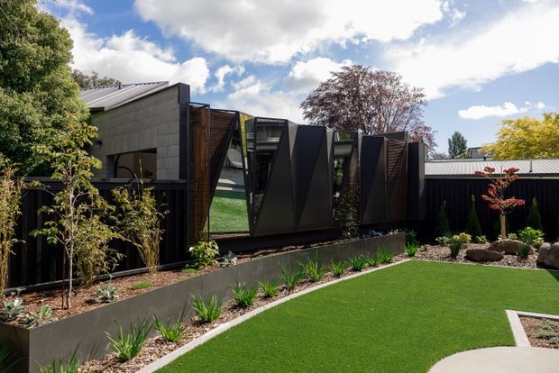 Contemporary single-storey building viewed from a landscaped courtyard, featuring dark vertical cladding, angular projecting window or screen elements, timber-toned slatted sections, and a low-pitched metal roof. The foreground has manicured lawn, planted garden beds, raised retaining edges, rocks, and a curved concrete path. Bright outdoor setting with a dramatic geometric facade.