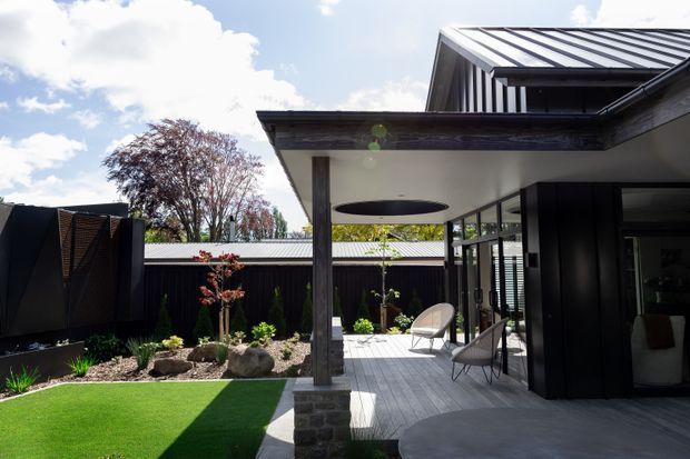 Contemporary single-storey residential courtyard viewed from the garden, with a covered outdoor terrace, dark metal-clad walls, a pitched metal roof, extensive glazing, timber-toned structural posts, and a landscaped garden with artificial turf, rocks, and planting. The terrace includes outdoor lounge chairs and a circular ceiling detail.