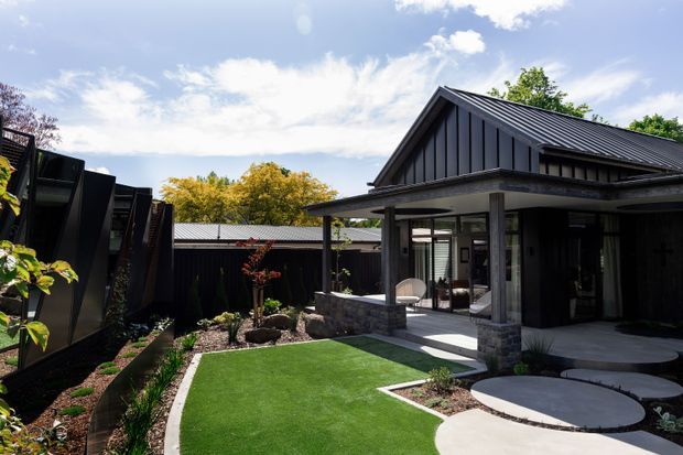 Exterior view of a contemporary dark-clad, gable-roofed residence opening onto a landscaped courtyard. The home features black vertical cladding, a covered veranda with square posts and stone-faced bases, extensive glazing, concrete paving, circular stepping pads, raised garden beds, and a small artificial-turf lawn.