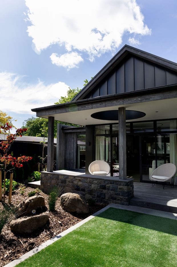 Exterior view of a contemporary single-storey gabled house with dark vertical metal cladding, a broad covered outdoor terrace, timber-like posts, large glazed doors, and a low natural-stone retaining wall. The landscaped foreground includes artificial turf, boulders, mulch, and planting.