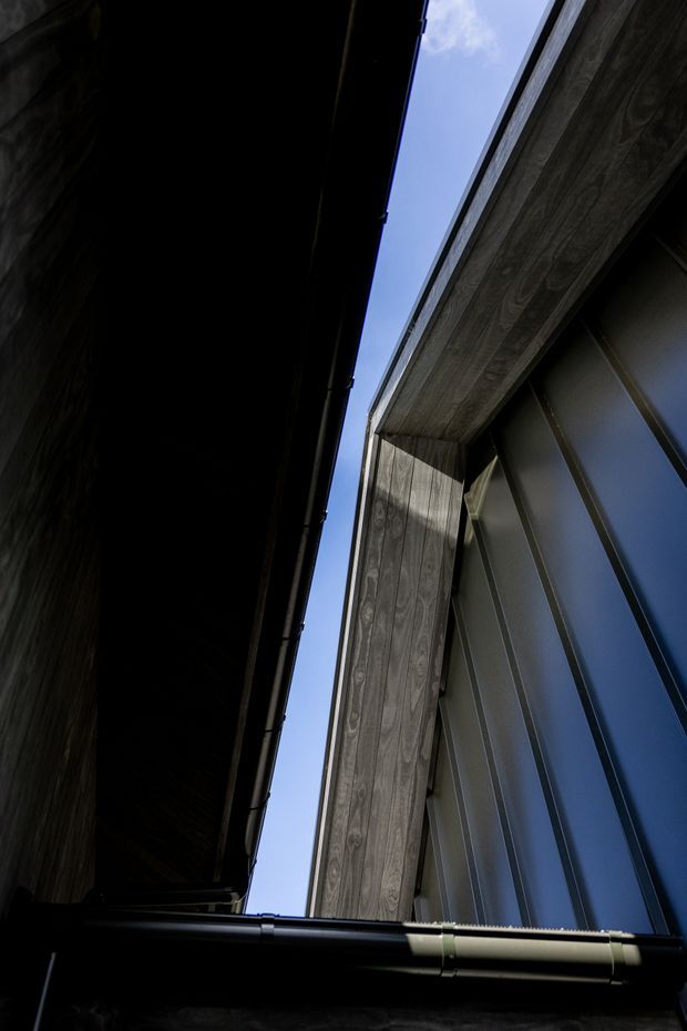 Upward-looking architectural detail between two closely spaced building forms. The image shows dark timber-clad walls and soffits, exposed timber framing, black metal gutters and downpipes, and a tall dark metal-clad wall with vertical seams and reflective glazing or panels. A narrow strip of blue sky is visible in the gap, creating a dramatic, high-contrast composition.