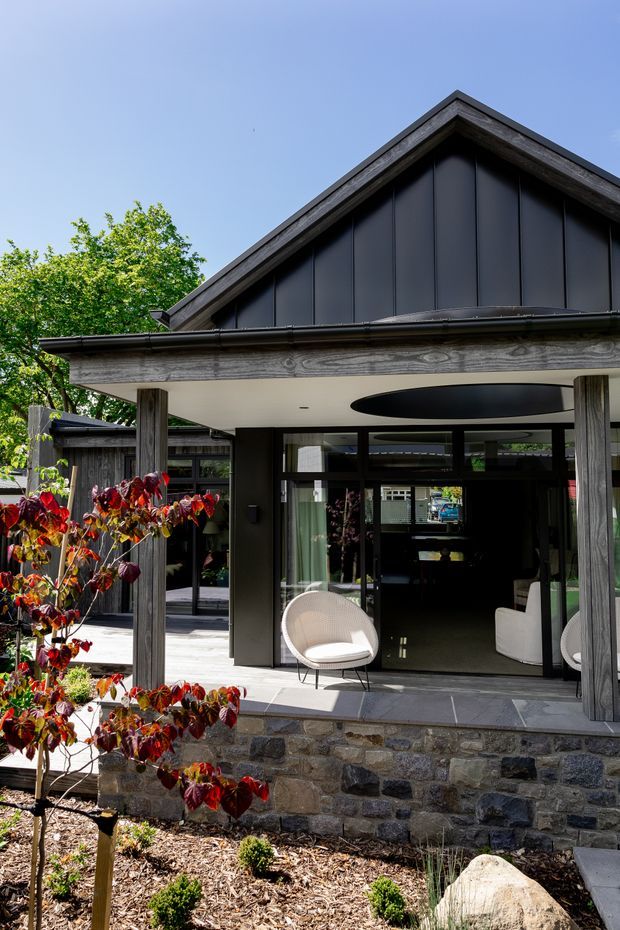 Exterior view of a contemporary gabled building with dark vertical cladding, exposed timber posts and beams, extensive glazed openings, and a covered outdoor seating area. The structure sits on a substantial rough stone base, with a landscaped garden in the foreground. A dark oval ceiling feature is visible beneath the covered entry or terrace.