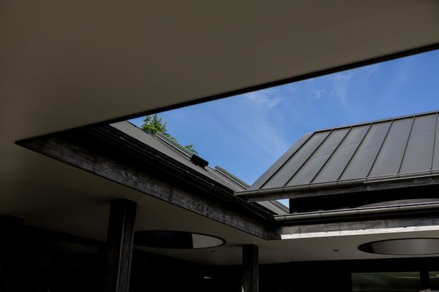 Architectural detail looking upward into a sheltered courtyard or covered passage, framed by dark timber or metal beams and roof edges. A bright blue sky is visible through the open central void, with contrasting pitched metal roofing and recessed circular ceiling details below.