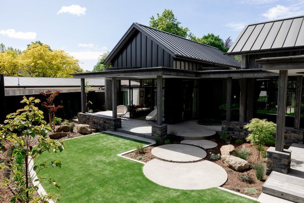 Exterior view of a contemporary single-storey home with dark standing-seam metal gable roofs, deep covered outdoor living areas, timber posts, extensive glazing, and a landscaped courtyard garden. Circular concrete stepping pads lead through artificial turf and planted beds toward the house.