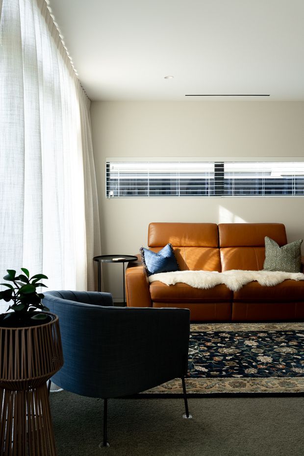 A bright, refined living room with a warm tan leather sofa, dark blue upholstered armchair, patterned area rug, small side table, indoor plant, and floor-to-ceiling sheer curtains. A long horizontal window with blinds brings in natural light, while a slim linear ceiling vent and recessed downlight contribute to the clean contemporary interior. The palette combines cream walls, brown leather, navy upholstery, and muted patterned textiles.
