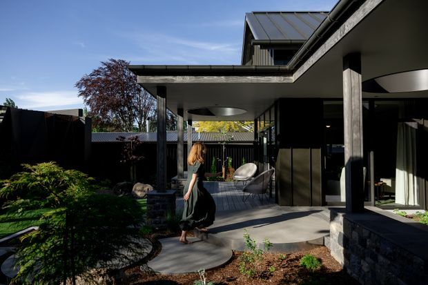Contemporary residential courtyard viewed from an outdoor garden path, with dark stone-clad walls, slender black structural columns, extensive glazing, and a covered terrace connecting the house around a landscaped courtyard. The low-pitched metal roof and layered flat-roof forms create a modern, sheltered outdoor living space.