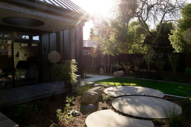 A landscaped residential courtyard beside a contemporary house, viewed from an outdoor deck. The garden features large irregular circular concrete stepping pads, low planting, ornamental grasses, rocks, and a narrow strip of lawn enclosed by timber fencing. The house has dark cladding, large glazed doors, a projecting roof with a visible metal edge, and a recessed oval ceiling light beneath the covered deck. Strong late-afternoon sunlight creates a warm, tranquil atmosphere.