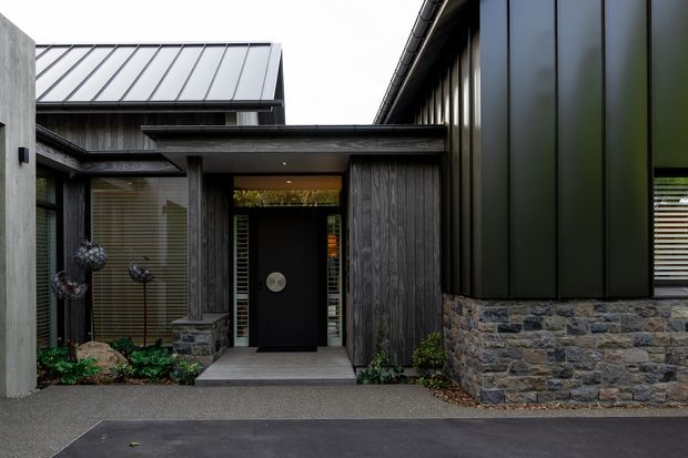 Contemporary residential entry facade featuring dark vertical timber cladding, charcoal metal standing-seam roofing, large dark metal wall panels, and a low natural stone masonry wall. A recessed front door is sheltered beneath a flat-roofed canopy, with narrow glazing and understated landscaping around the entrance.
