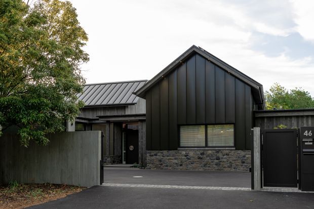 Front exterior view of a contemporary single-storey dwelling with a steep gabled roof, dark vertical metal cladding, a low stone-clad wall, and a recessed entry behind a dark boundary fence and gate. The facade is framed by mature landscaping and includes horizontal louvred window blinds.