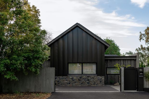 Front exterior view of a compact contemporary gabled house, featuring dark vertical metal cladding, a contrasting low stone masonry base, timber fencing, and a gated entry integrated into the street frontage. Mature trees partially frame the property.