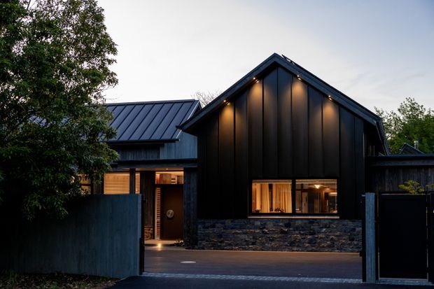 Exterior view of a contemporary dark-clad house at dusk, seen through an open driveway gate. The home has steep gabled roof forms, vertical black metal cladding, a low wall of dark rough stone, warm-lit timber-framed windows, and integrated exterior lighting. Mature planting partially screens the left side.