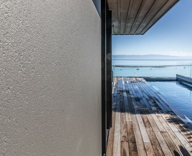 A close architectural view from a covered outdoor area, showing a textured pale plaster wall and dark framed opening leading onto a weathered timber deck. An infinity-edge pool occupies the right side, with expansive blue water and distant mountains beyond. The overhead soffit features timber boards, creating a strong indoor–outdoor connection and a serene waterfront setting.