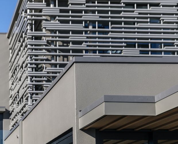 Close-up exterior view of a contemporary multi-storey building. A lower rendered volume with dark metal trim and a projecting canopy sits in front of a taller glazed facade screened by dense horizontal metal sun-shading fins. The composition emphasizes layered geometry, modern facade detailing, and strong linear shadows against a clear blue sky.