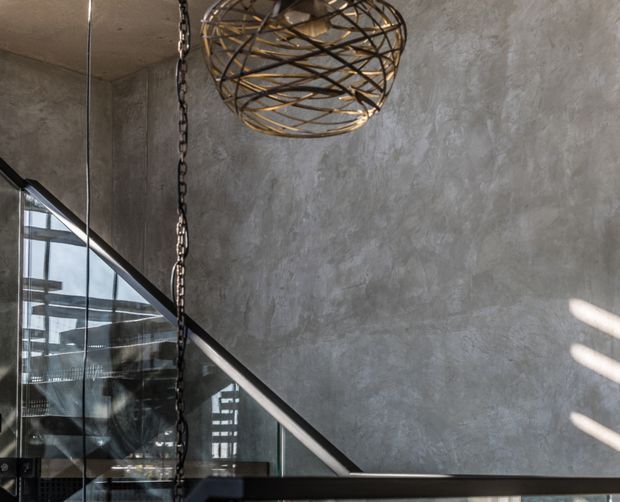 A close architectural interior view of a modern stairway or landing with a dark metal and glass balustrade, a large textured grey concrete-like wall, and an oversized spherical woven metal pendant light suspended from a chain. Strong sunlight creates linear highlights on the wall, while glazing reveals additional stair or structural elements beyond.