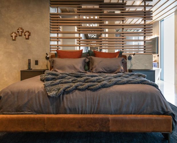 A warmly styled bedroom centered on a large bed with a dark taupe upholstered headboard and matching bedding. Layered grey and rust-orange cushions, a chunky knitted throw, dark bedside tables, and wall-mounted reading lights create a cozy, refined atmosphere. A horizontal timber slat screen separates the bed from a brighter adjoining area, while the exposed ceiling structure and light-filled glazing add architectural interest.