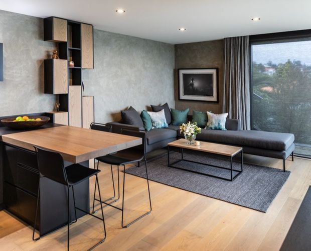 A contemporary open-plan living and dining area with a dark L-shaped sectional sofa, timber dining table and black chairs, floating timber cabinetry, textured grey walls, and wide floor-to-ceiling glazing with curtains. Light timber flooring, a charcoal rug, recessed ceiling lights, and muted blue-green cushions create a refined, warm modern interior.