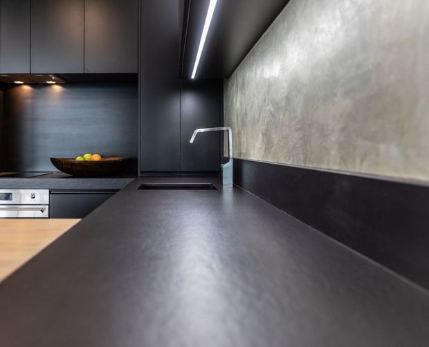 Close-up view of a sleek, dark contemporary kitchen featuring a broad matte charcoal benchtop, integrated black cabinetry, a dark splashback, undermount sink with angular chrome tapware, and warm timber flooring at the edge of the frame. A textured grey concrete-like wall finish and linear recessed lighting add an industrial, minimalist character.