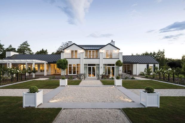 Front-facing exterior view of a large symmetrical white weatherboard residence with a dark pitched metal roof, central two-storey gabled section, broad covered verandah, and extensive glazed doors and windows. The formal landscaped forecourt features gravel paths, clipped lawns, raised white planters, low fencing, and established planting. The image is photographed at dusk under a pale blue sky.