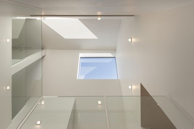 Upward-looking interior view of a minimalist stairwell or landing beneath a sloped ceiling, with a large skylight, a rectangular window, glass balustrades, white walls, and small recessed or wall-mounted lights. The composition emphasizes clean geometric forms, abundant daylight, and reflective glazing.