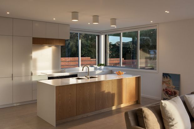 A bright, contemporary open-plan kitchen viewed from the adjacent living area. The room features a large timber-fronted island with an integrated sink and gooseneck tap, pale stone benchtops, full-height light cabinetry, a stainless-steel cooking zone, and expansive corner glazing overlooking a garden and brick boundary wall. Minimal recessed and cylindrical pendant lighting create a clean, warm interior.