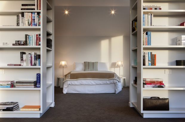 A symmetrical view through two tall built-in bookcases into a compact bedroom. The room contains a neatly made bed with layered neutral bedding, two bedside lamps, recessed ceiling spotlights, pale walls, and dark carpet. The open shelving is filled with books and a few storage objects, creating a calm, refined and highly organised interior.