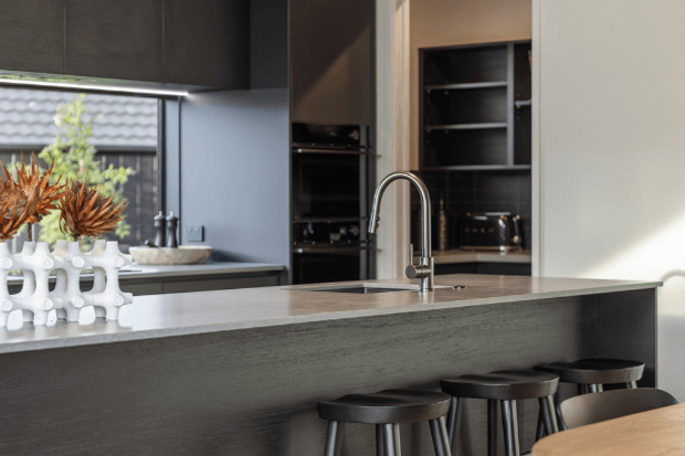 Contemporary kitchen with a large dark island, pale stone benchtop, integrated cabinetry and appliances, a tall curved chrome tap, and black stools. A dining table is partially visible in the foreground, while daylight enters through the window and reflects across the work surfaces.