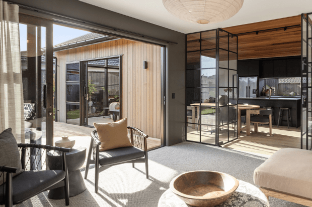 Sunlit contemporary open-plan living and dining area with a visible kitchen beyond. The room features floor-to-ceiling glazing, dark framed glass partitions, light textured carpeting, timber-lined ceiling and cabinetry, and a neutral palette with black metal accents. An outdoor courtyard and adjacent timber-clad structure are visible through the glazing.