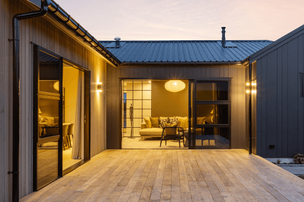 A warm dusk view of a private courtyard enclosed by timber-clad wings with dark standing-seam metal roofs. Wide timber decking leads to full-height glazed doors and windows, revealing a warmly lit living area with a sofa, pendant light and interior furnishings. Exterior wall lights, exposed gutters and downpipes, and dark metal cladding define the contemporary courtyard architecture.