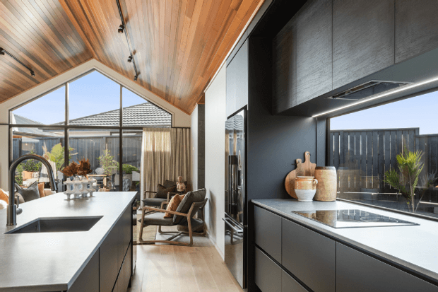 Contemporary open-plan kitchen with a dramatic pitched timber-lined ceiling, expansive glazing, and a large central island. Matte dark cabinetry and integrated appliances contrast with light stone-look benchtops, while a compact seating nook and landscaped courtyard are visible beyond the glazing.