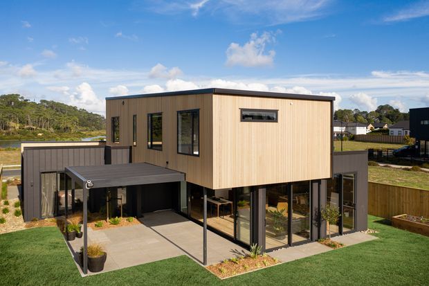 A modern two-storey residential building photographed from the exterior. The upper volume is clad in vertical light timber boards with dark-framed windows, while the lower level combines dark cladding, extensive glazing, and a covered outdoor patio or entry area. The house sits on a landscaped suburban site with lawn, garden beds, fencing, and a nearby water edge visible in the background.