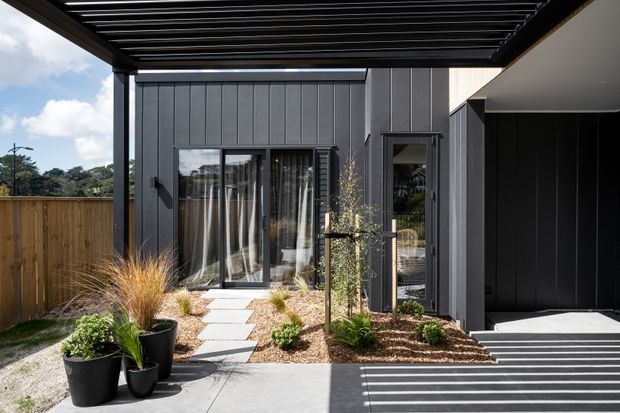 Contemporary dark-clad residential exterior with vertical panel cladding, a covered slatted roof structure, and a landscaped entry courtyard. A paved stepping-stone path leads through gravel planting beds to glazed doors, while a covered carport or entry canopy occupies the right side.