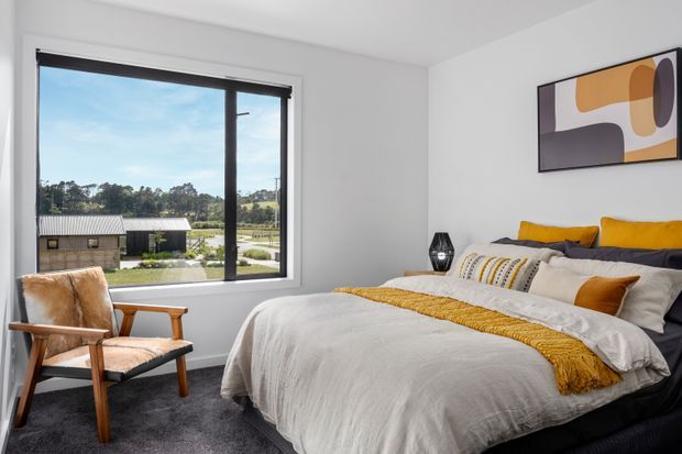 Bright, contemporary bedroom with a large black-framed window overlooking a rural landscape. The room features a low dark upholstered bed, white textured bedding with mustard-yellow accents, a dark carpet, a mid-century-style timber and upholstered lounge chair, black bedside lighting, and abstract geometric wall art.