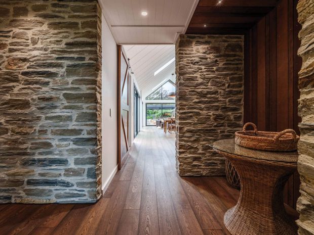 Interior view down a long, refined hallway framed by substantial exposed natural-stone walls. The space has warm timber flooring, white tongue-and-groove ceiling panels, dark timber detailing and doors, and a glazed opening at the far end leading toward a bright living or dining area. A woven rattan console and basket add natural texture in the foreground.