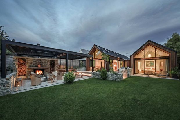 A low-rise residential compound arranged around a broad lawn and outdoor entertaining courtyard. Several dark-clad, steep-gabled pavilion forms are linked by glazed passages and framed with warm timber. The left pavilion contains an open-air or semi-enclosed fireplace lounge with a stacked stone wall and timber storage, while the central and right pavilions show warmly lit interiors through extensive glazing. The composition has a refined contemporary lodge or cabin character, with stone retaining and paving, landscaped planting, and a dramatic overcast evening sky.