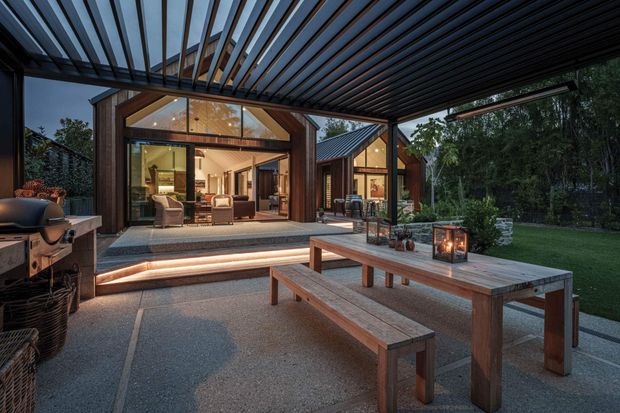 Evening view of a covered outdoor entertaining area with a timber picnic-style dining table and benches, barbecue, lanterns, and a small fire feature. The space opens directly onto a warm, timber-clad interior through large glazed doors, with illuminated steps and a lush garden beyond. A slatted overhead canopy creates a sheltered, architectural outdoor room.
