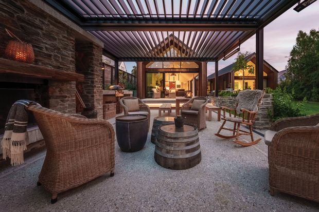 A covered outdoor entertaining terrace with a slatted pergola roof, stone fireplace wall, gravel floor, and woven rattan lounge furniture arranged around barrel-style coffee tables. A glazed living area and timber-clad building wings are visible beyond, with landscaped planting at the edges. The scene has a warm, rustic lodge-like atmosphere at dusk.